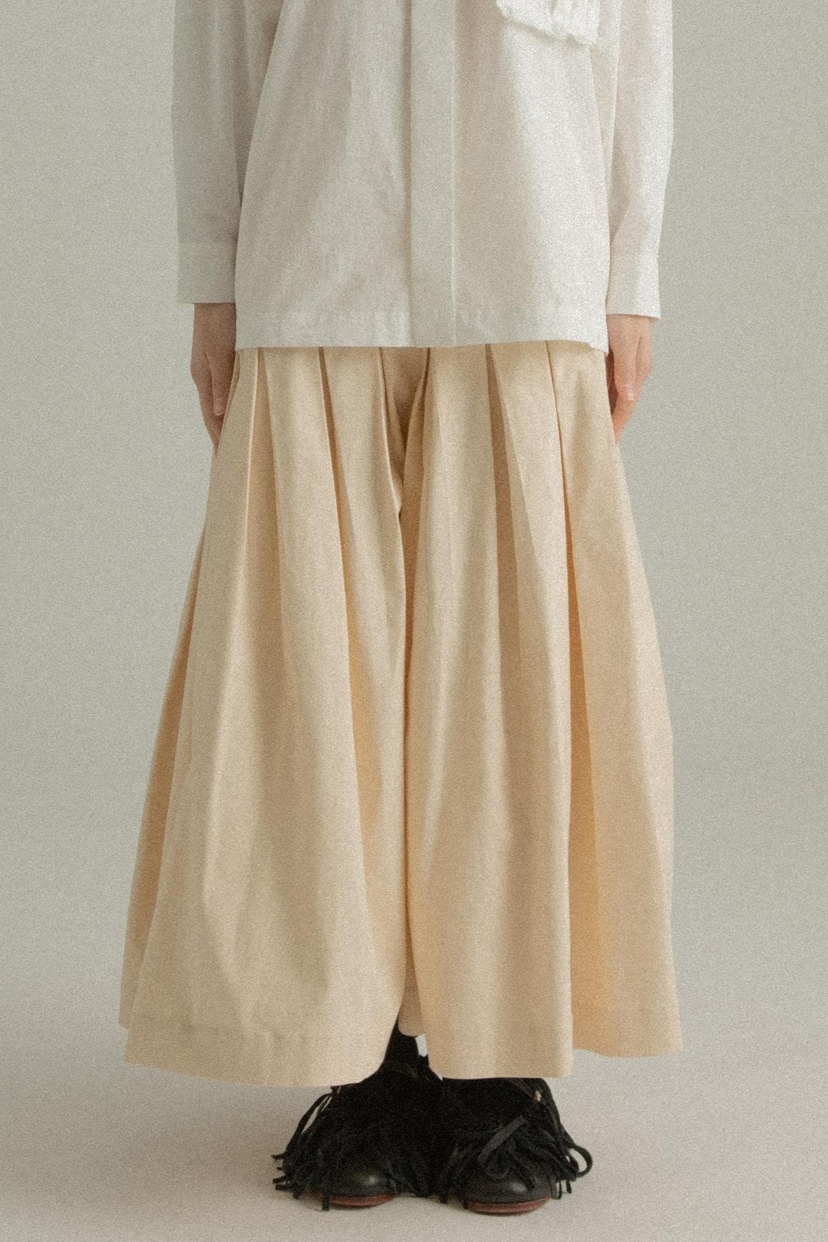 A model wearing the Clara Skirt in a soft A-line silhouette with diagonal pleating. The skirt is made from a Tencel and linen blend, styled with the Charlotte Shirt. The model is shown in front, side, and back views.