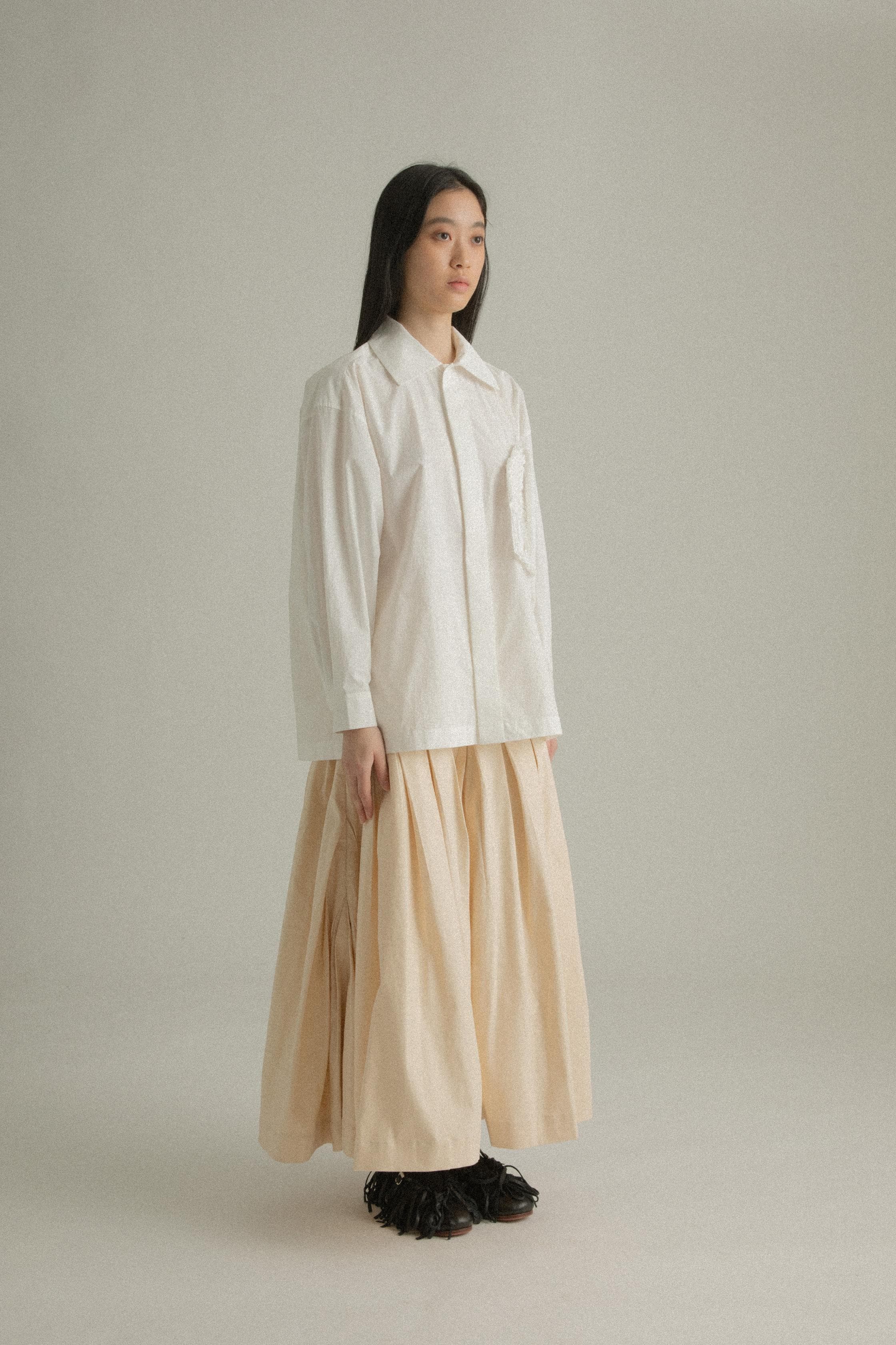 A model wearing the Clara Skirt in a soft A-line silhouette with diagonal pleating. The skirt is made from a Tencel and linen blend, styled with the Charlotte Shirt. The model is shown in front, side, and back views.