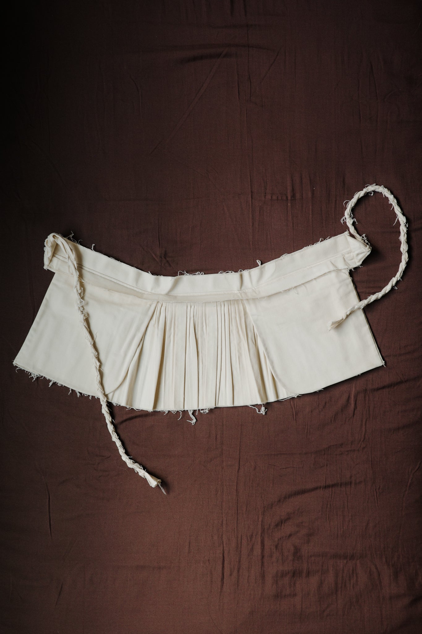 Cream pleated Goma apron with structured waistband and braided ties.