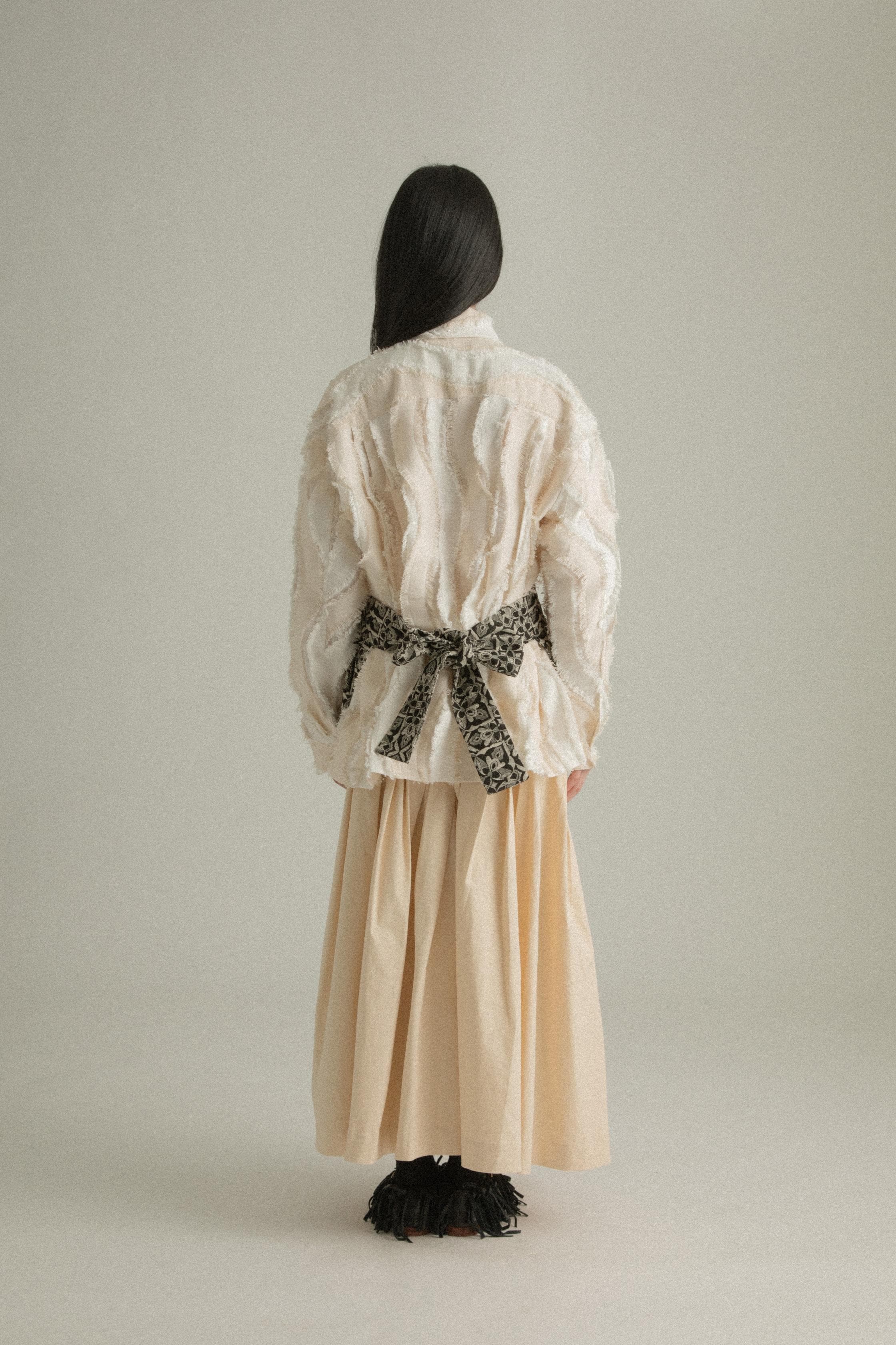 A model wearing the Percy Shirt in cream, made from Chinese cotton jacquard with a unique textural pattern. The shirt has a classic silhouette with a structured collar and concealed button placket. It is styled with the Delia Apron, which is not part of the shirt. The model is shown in front, side, and back views.