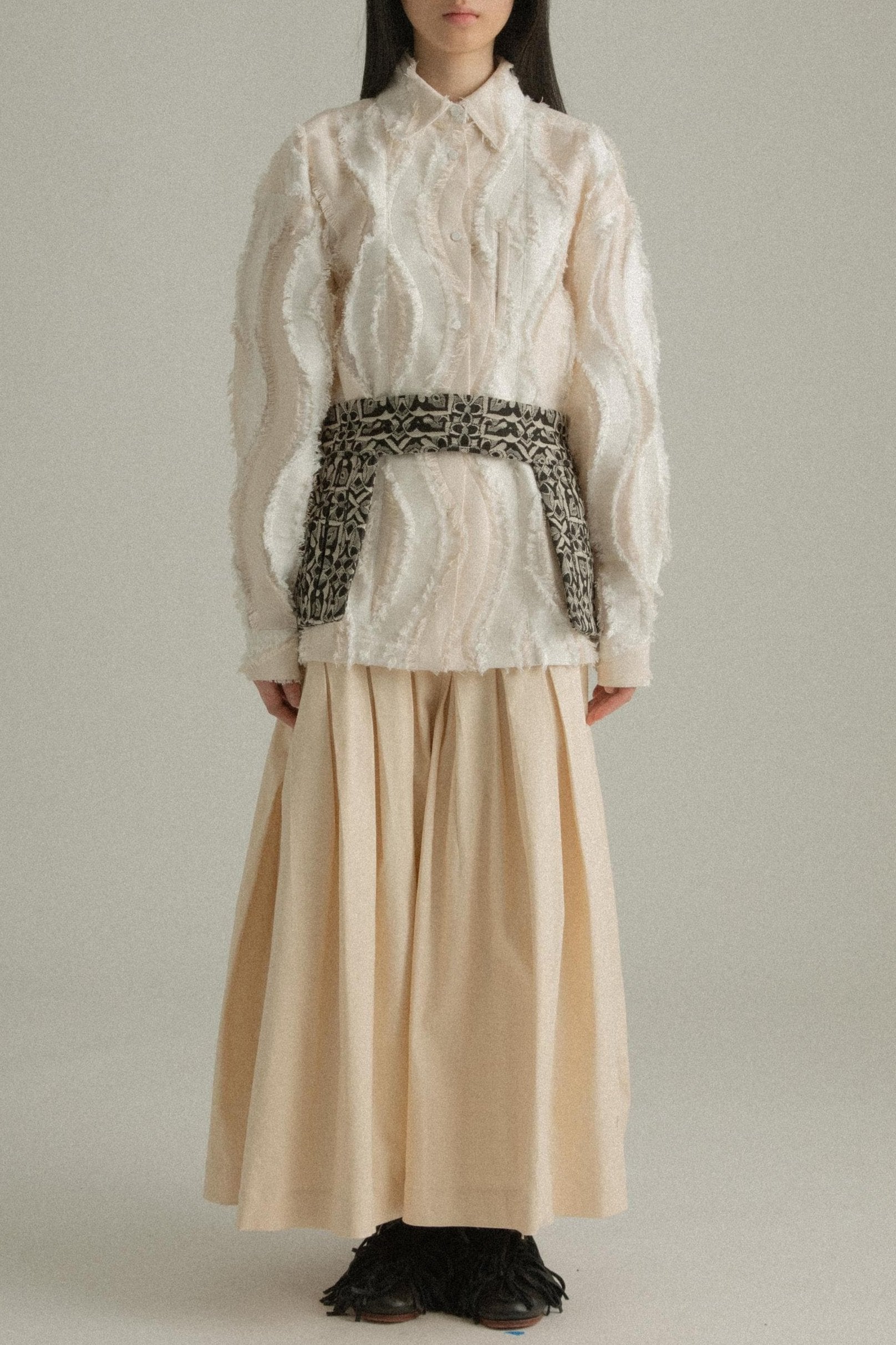 A model wearing the Percy Shirt in cream, made from Chinese cotton jacquard with a unique textural pattern. The shirt has a classic silhouette with a structured collar and concealed button placket. It is styled with the Delia Apron, which is not part of the shirt. The model is shown in front, side, and back views.