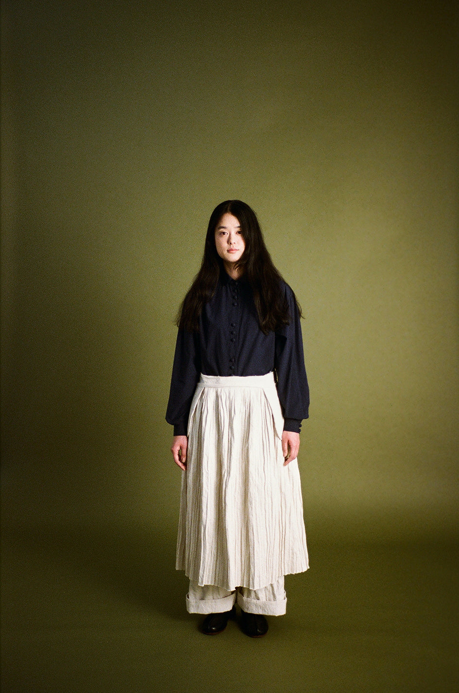 A woman stands against a solid olive green studio backdrop wearing a dark navy long-sleeve blouse and a cream pleated midi skirt.