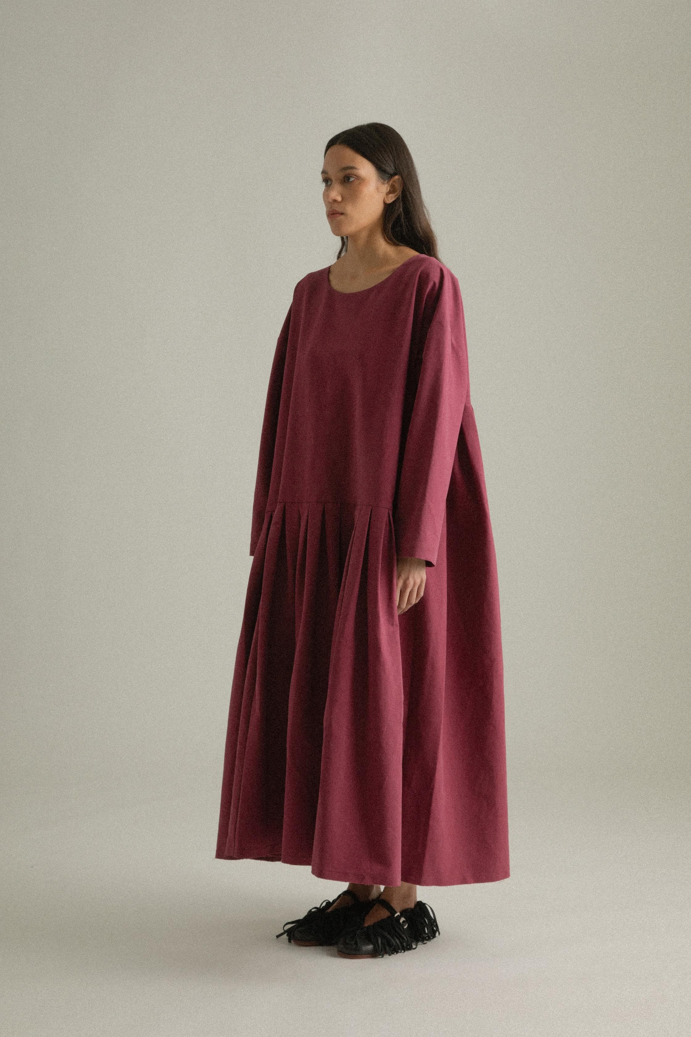 A model wearing the Arya Dress, crafted from 100% bony cotton with a loose, asymmetric round neckline and ankle-length gathered pleats. The dress can be worn front-to-back or back-to-front and features side pockets. The model is shown in front, side, and back views.