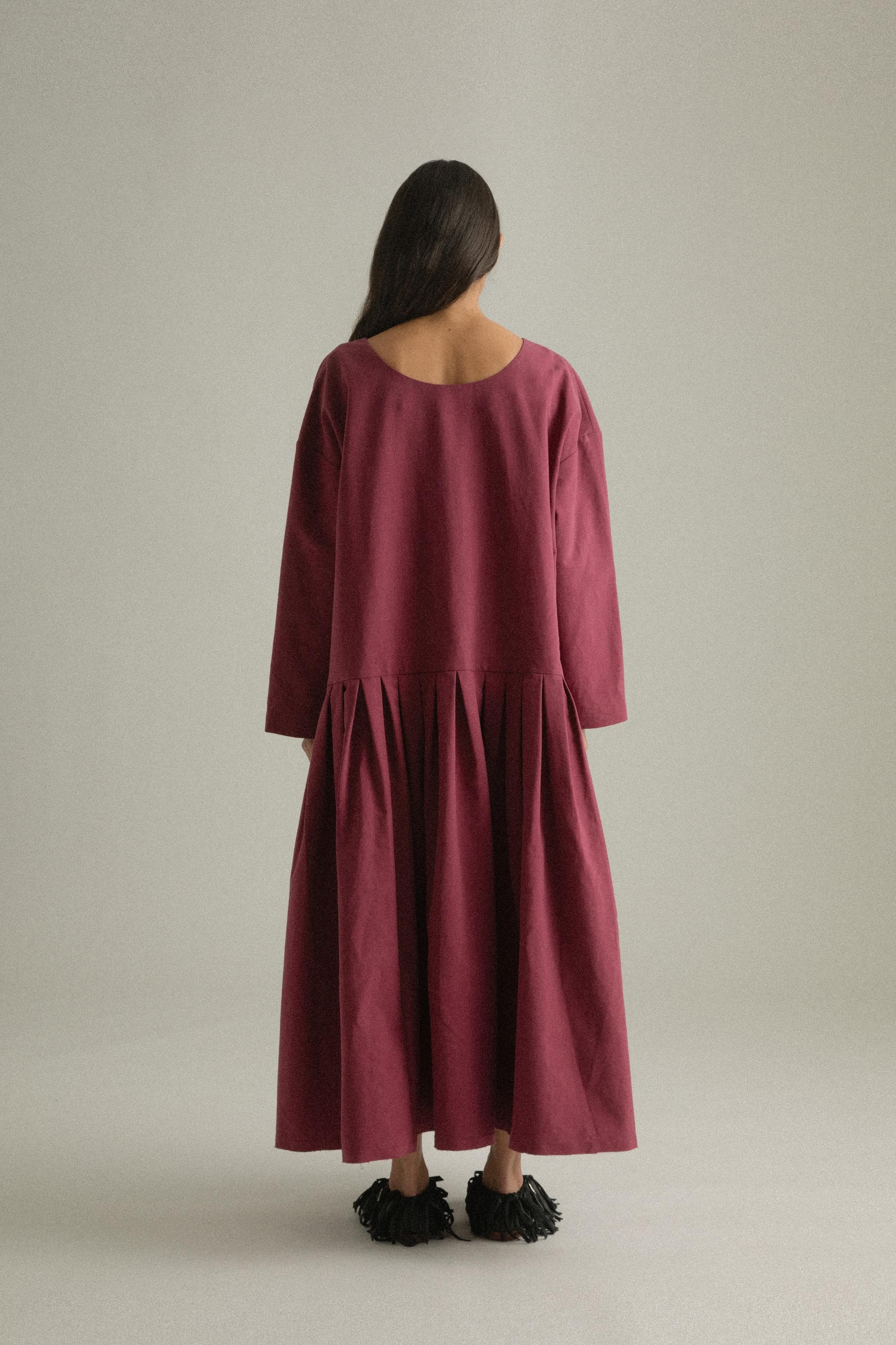 A model wearing the Arya Dress, crafted from 100% bony cotton with a loose, asymmetric round neckline and ankle-length gathered pleats. The dress can be worn front-to-back or back-to-front and features side pockets. The model is shown in front, side, and back views.