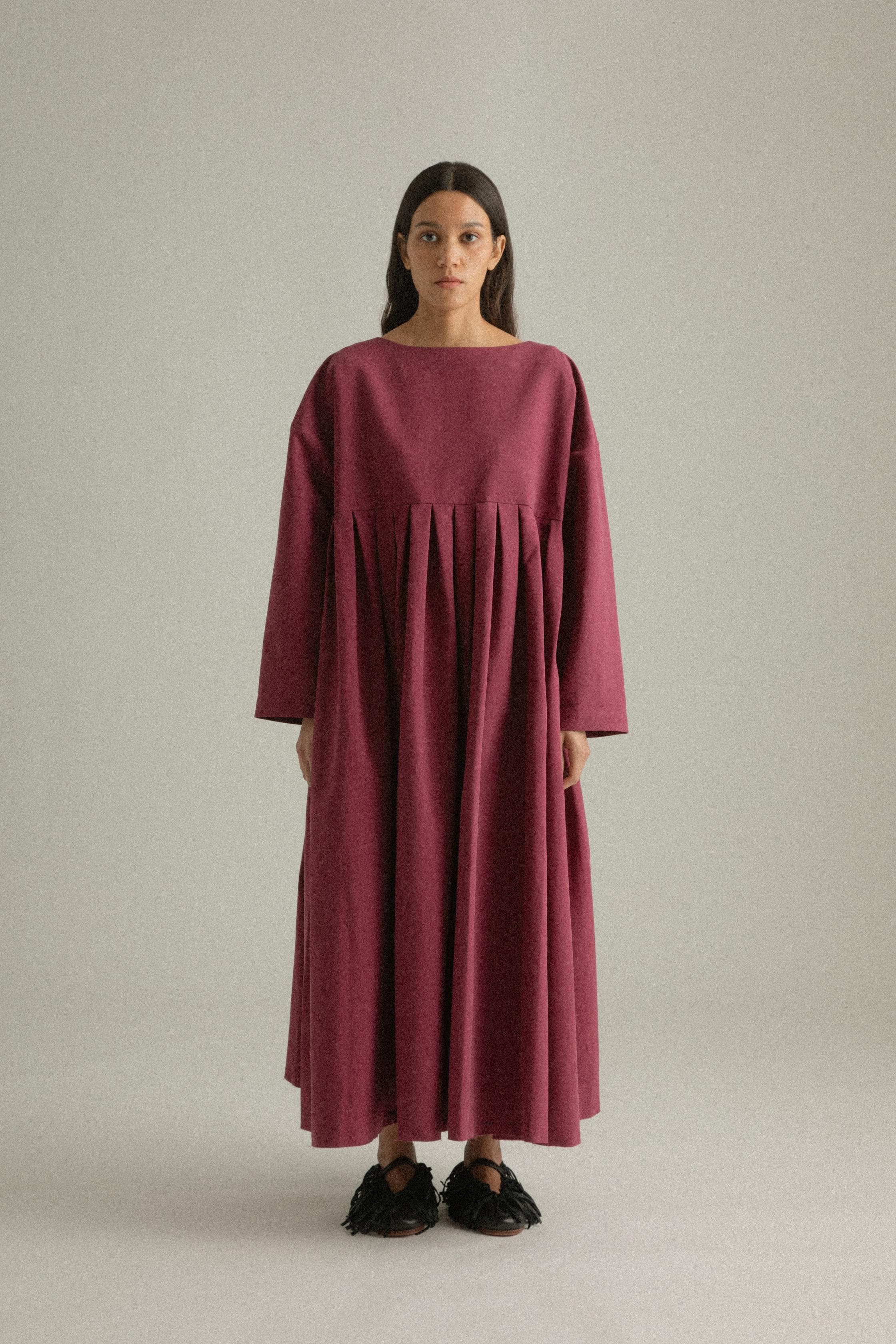 A model wearing the Arya Dress, crafted from 100% bony cotton with a loose, asymmetric round neckline and ankle-length gathered pleats. The dress can be worn front-to-back or back-to-front and features side pockets. The model is shown in front, side, and back views.
