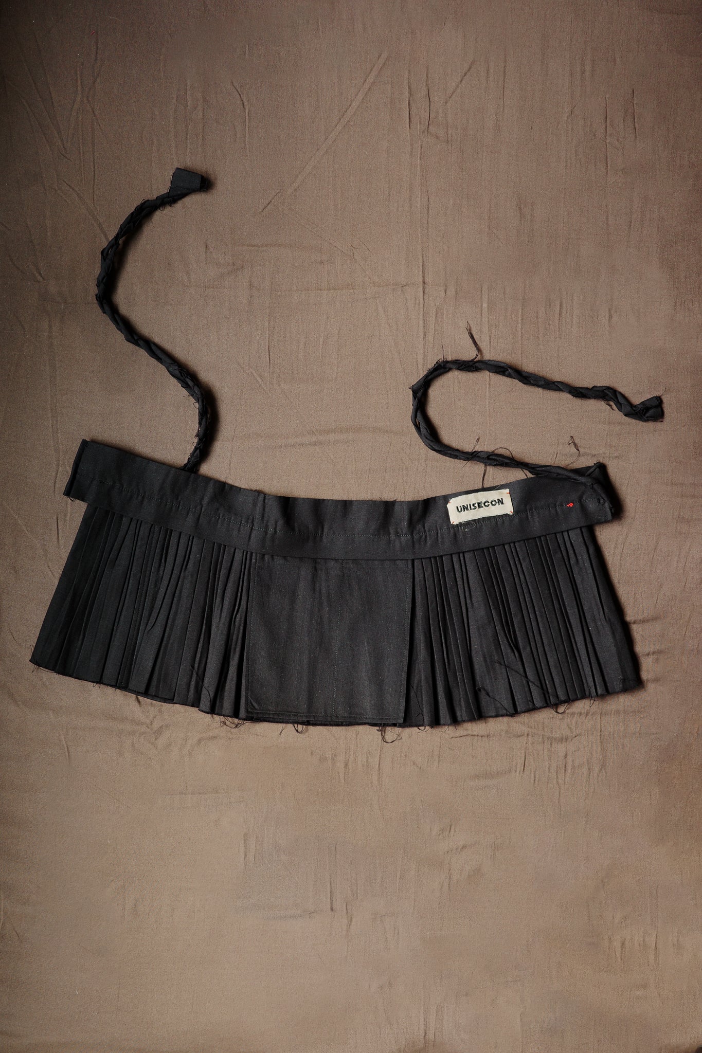 Black pleated Goma apron with braided ties and a UNISECON label.