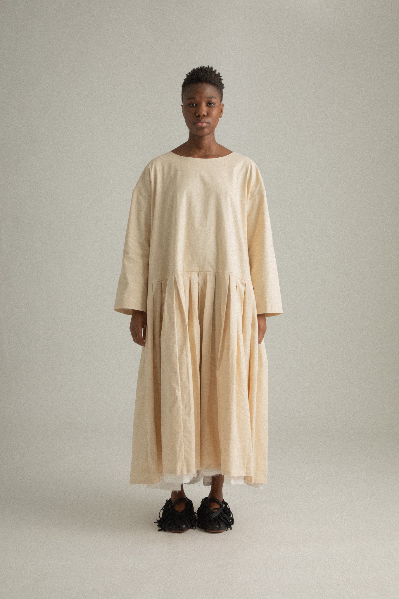 Front view of Arya Dress in compact cream cotton, showcasing the curved neckline and pleated skirt.