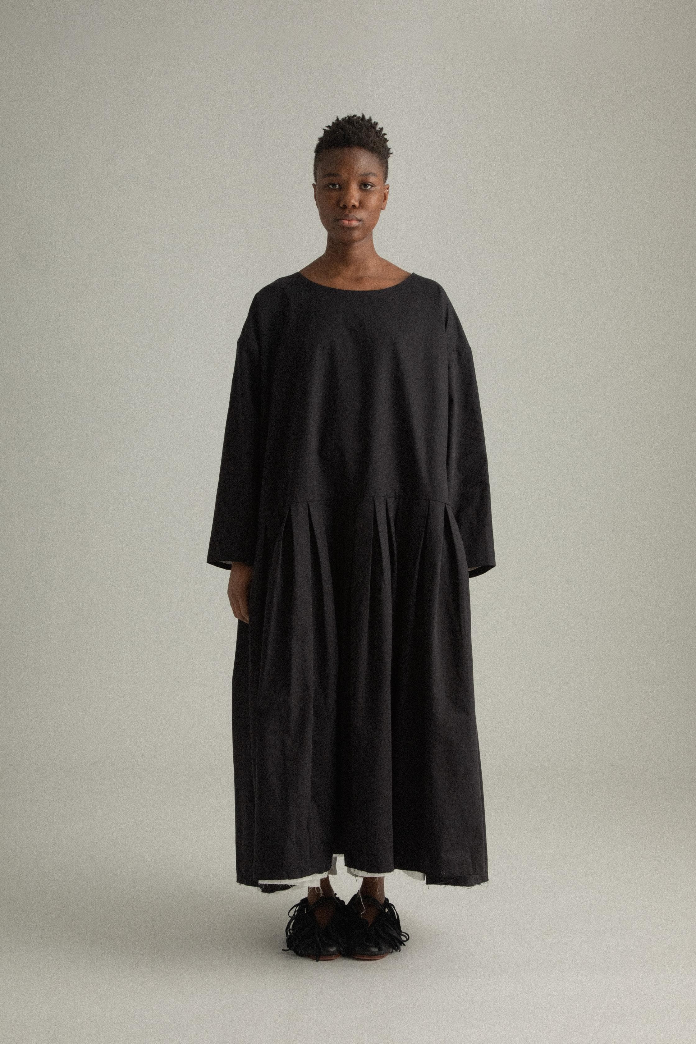 A model wearing the Arya Dress, a loose-fitting, ankle-length dress made of 100% organic cotton. The dress features an asymmetrical neckline, subtle pleating, and a relaxed silhouette. It is designed for reversible wear and has side pockets. The inner white lining is slightly visible at the hem, adding a minimalist contrast to the black fabric. The model is shown in front, side, and back views to highlight the dress’s design and fit.