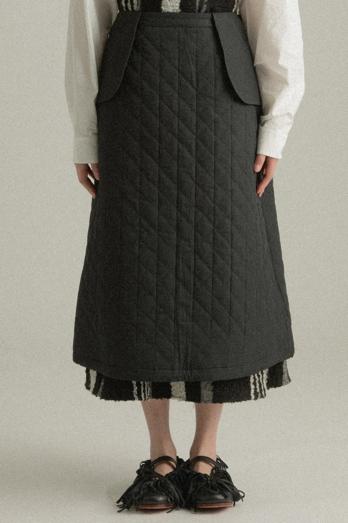 A model wearing the Lulu Skirt, crafted from 100% organic cotton with a quilted texture. The skirt features an A-line silhouette and is layered over the Lena Dress, adding dimension and warmth. The model is shown in front, side, and back views.