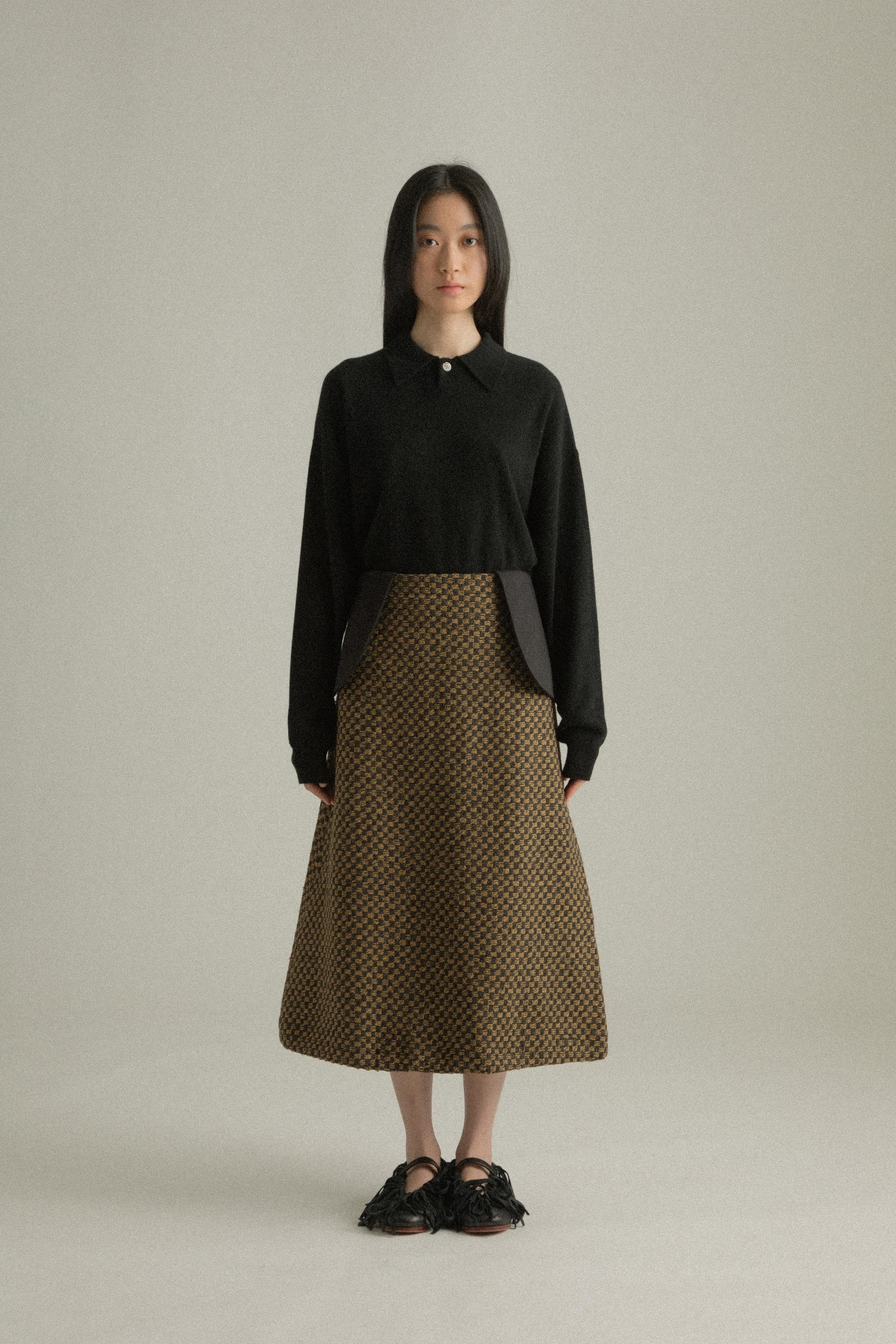 A model wearing the Lulu Skirt, featuring a classic A-line silhouette crafted from a blend of wool and cotton with a jacquard pattern. The skirt showcases a checkered design in black and golden hues. The model is shown in front, side, and back views.