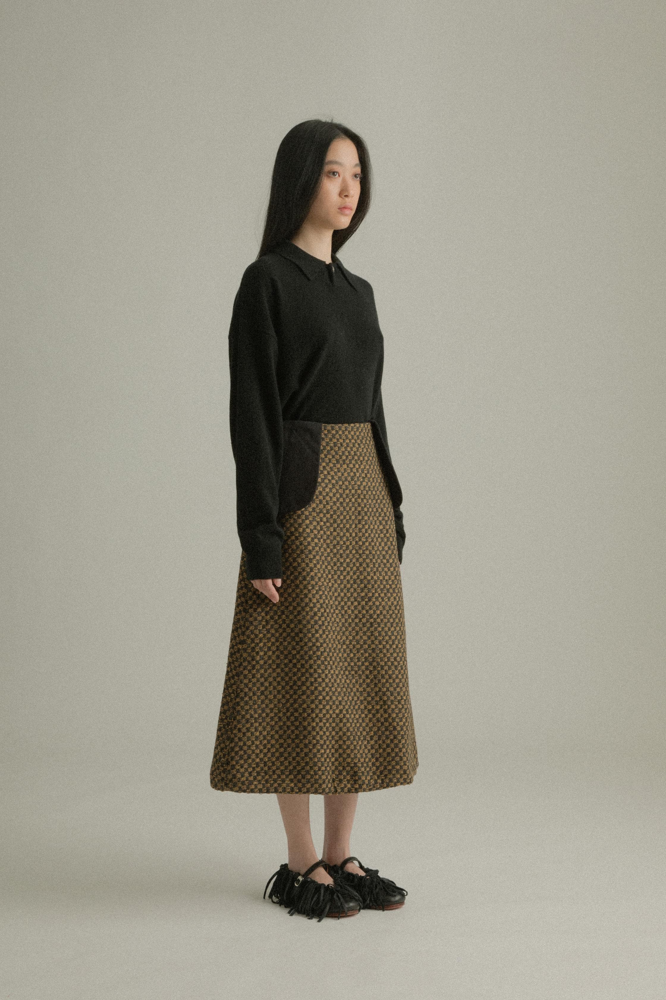 A model wearing the Lulu Skirt, featuring a classic A-line silhouette crafted from a blend of wool and cotton with a jacquard pattern. The skirt showcases a checkered design in black and golden hues. The model is shown in front, side, and back views.