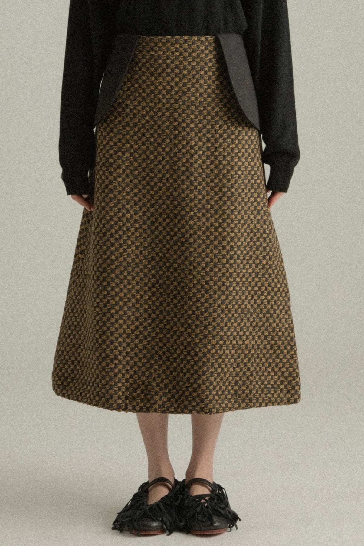 A model wearing the Lulu Skirt, featuring a classic A-line silhouette crafted from a blend of wool and cotton with a jacquard pattern. The skirt showcases a checkered design in black and golden hues. The model is shown in front, side, and back views.