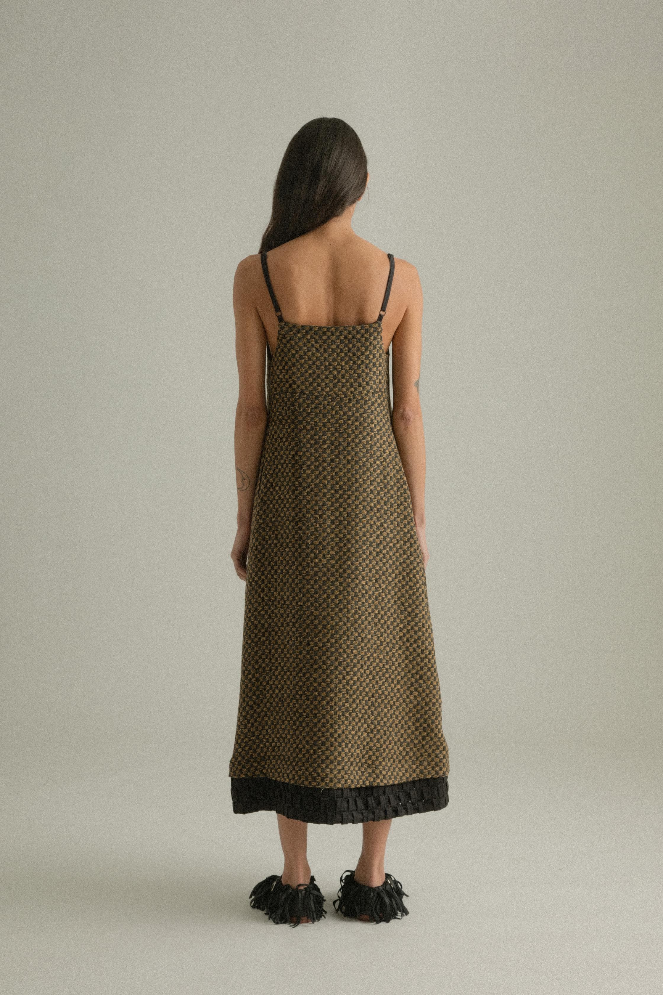 A model wearing the Mila Dress, crafted from a blend of wool and cotton with an intricately handwoven jacquard pattern in black and golden hues. The dress features a straight silhouette with delicate straps and a detailed hem. The model is shown in front, side, and back views.