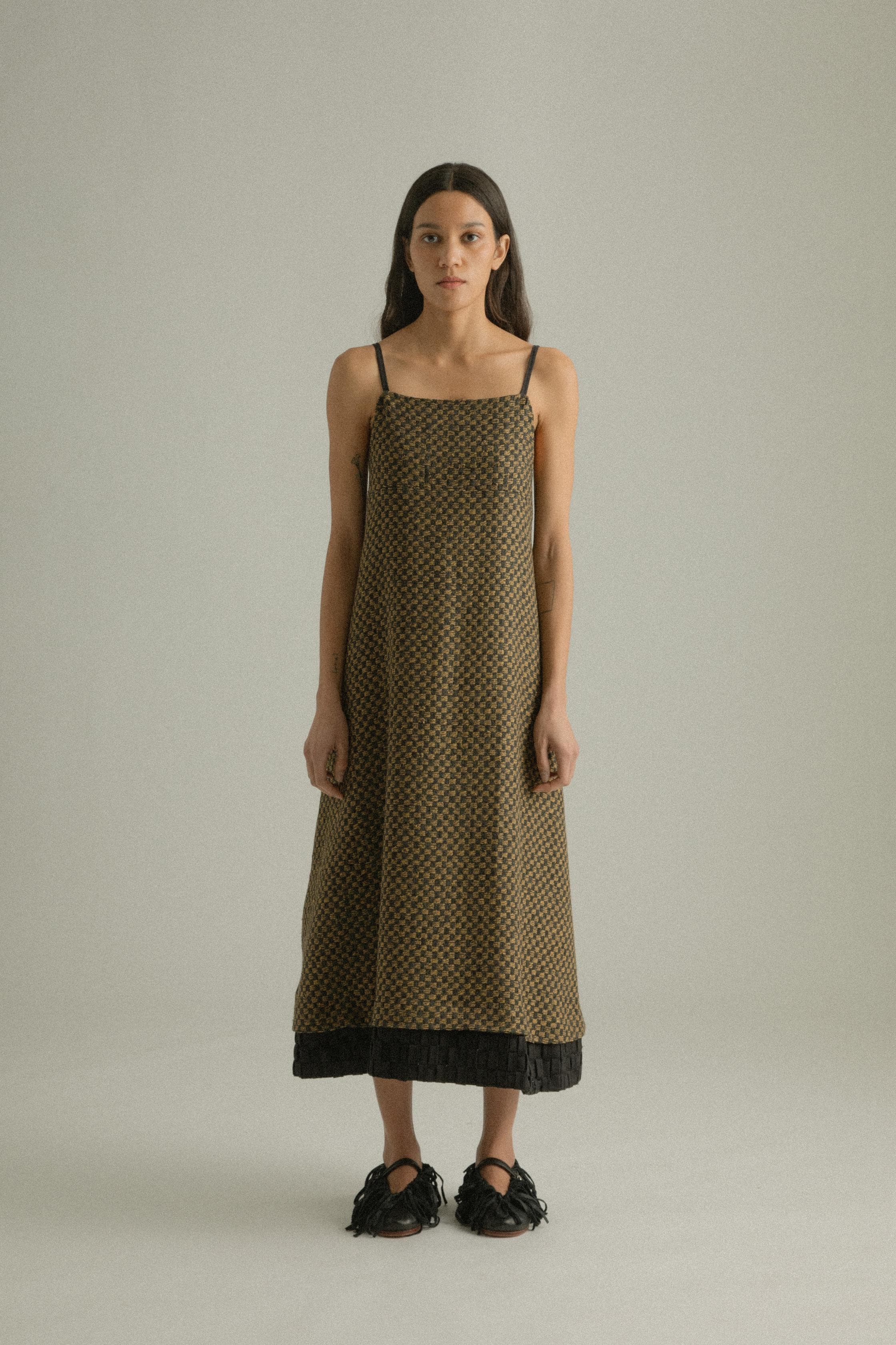 A model wearing the Mila Dress, crafted from a blend of wool and cotton with an intricately handwoven jacquard pattern in black and golden hues. The dress features a straight silhouette with delicate straps and a detailed hem. The model is shown in front, side, and back views.