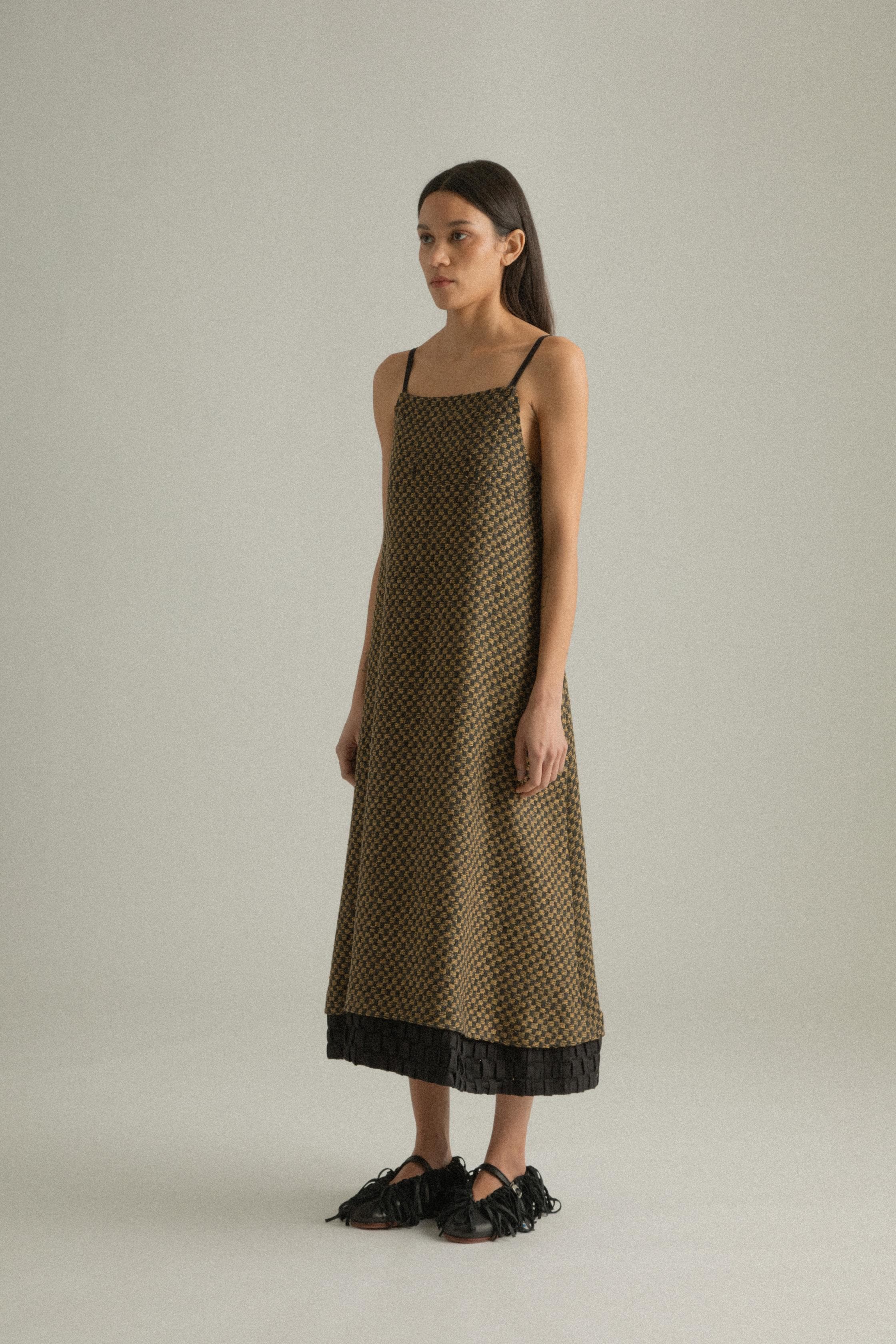 A model wearing the Mila Dress, crafted from a blend of wool and cotton with an intricately handwoven jacquard pattern in black and golden hues. The dress features a straight silhouette with delicate straps and a detailed hem. The model is shown in front, side, and back views.