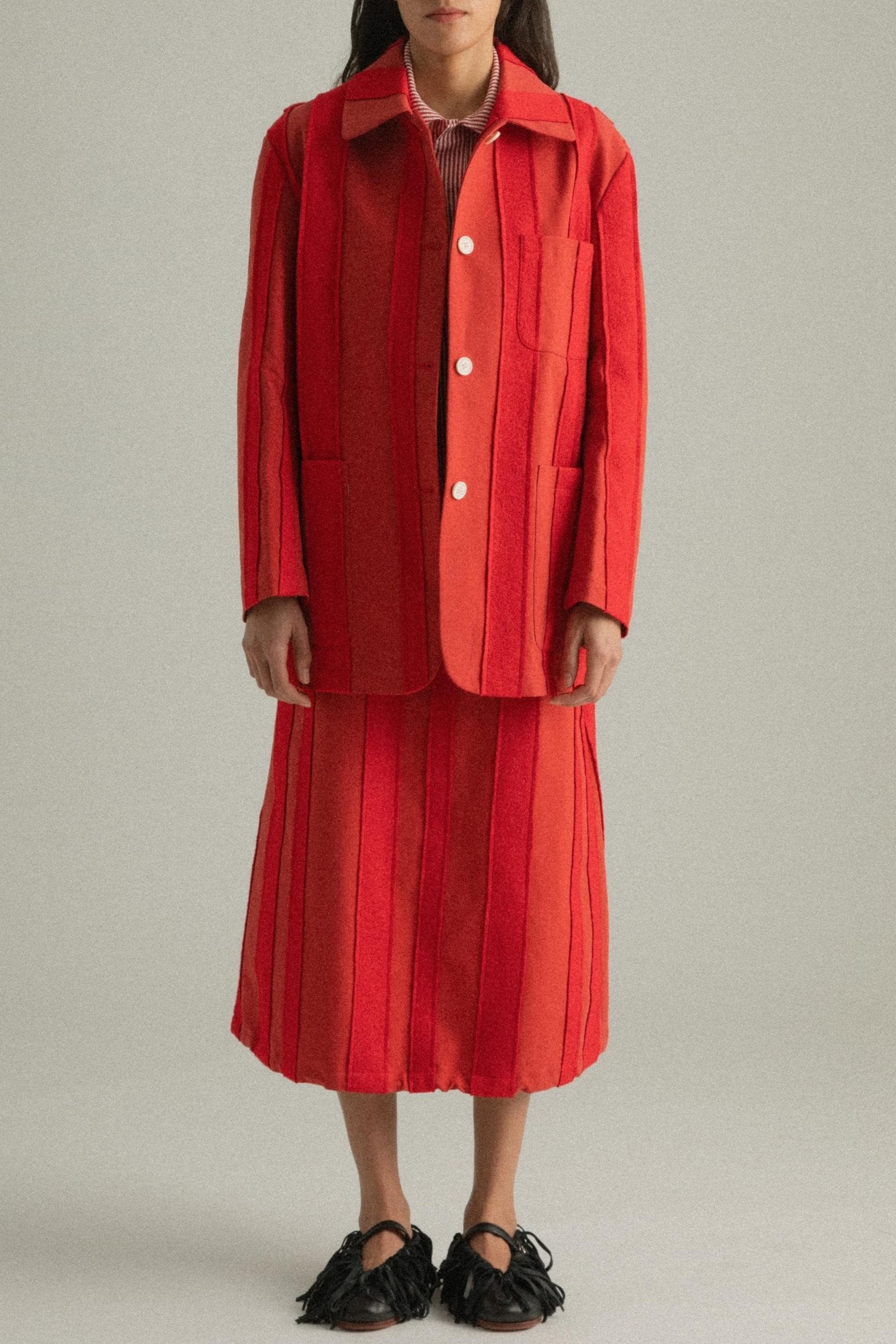 A model wearing the Nicole Jacket in a bold red hue, crafted from 100% cotton with intricate hand-stitching. The jacket features a round collar and single-button closure, styled with the Lulu Skirt. The model is shown in front, side, and back views.