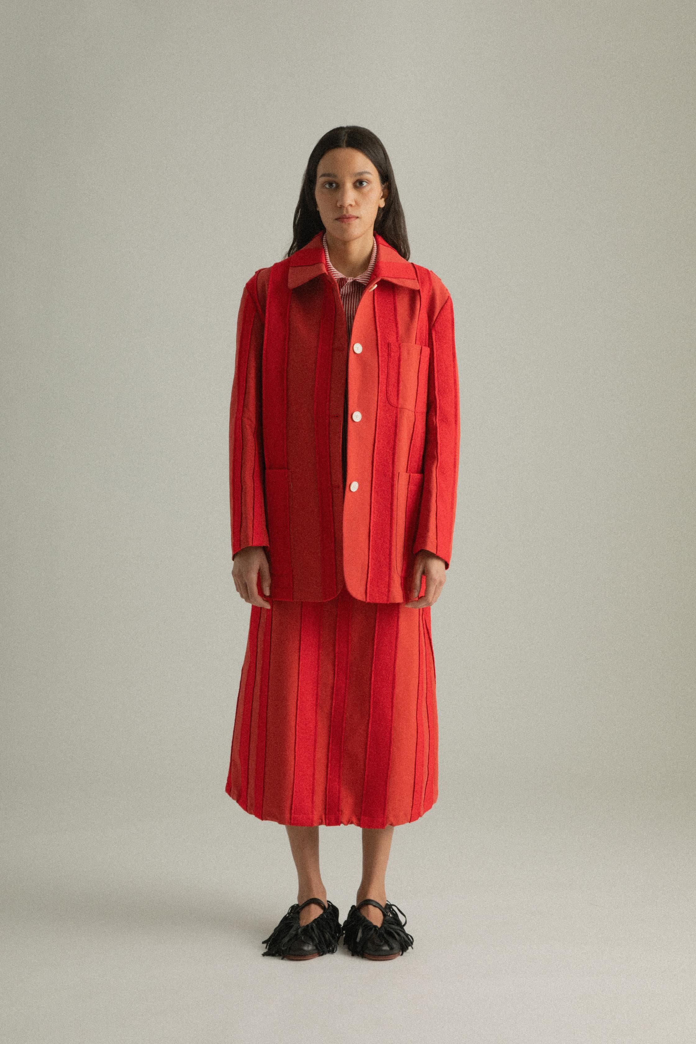 A model wearing the Nicole Jacket in a bold red hue, crafted from 100% cotton with intricate hand-stitching. The jacket features a round collar and single-button closure, styled with the Lulu Skirt. The model is shown in front, side, and back views.