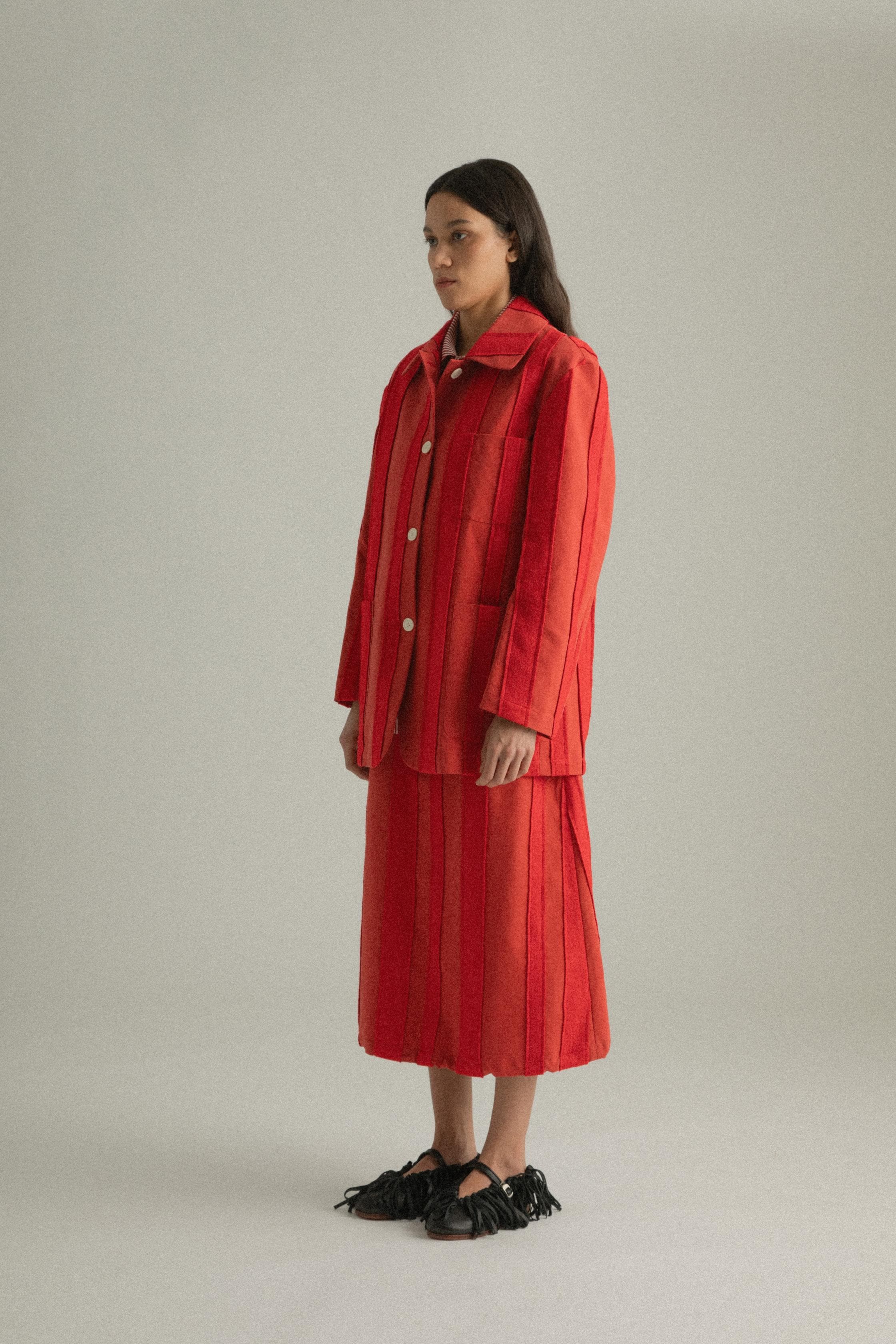 A model wearing the Nicole Jacket in a bold red hue, crafted from 100% cotton with intricate hand-stitching. The jacket features a round collar and single-button closure, styled with the Lulu Skirt. The model is shown in front, side, and back views.