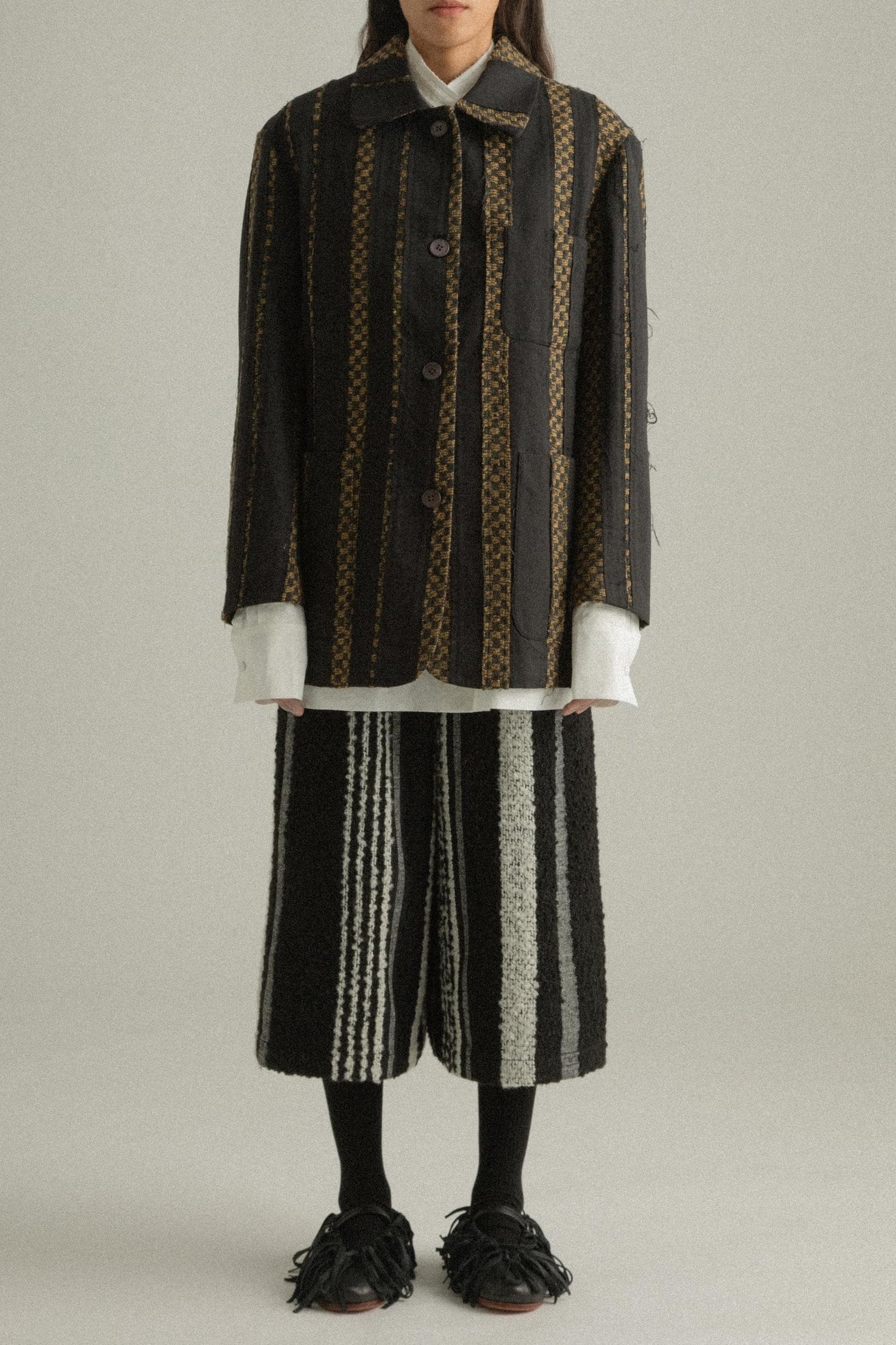 A model wearing the Nicole Jacket, featuring a classic round collar and single-button closure. The jacket is crafted from handwoven fabric in a blend of wool, cotton, and polyester with black and gold hues. Styled with DI Pants for a cohesive look. The model is shown in front, side, and back views.
