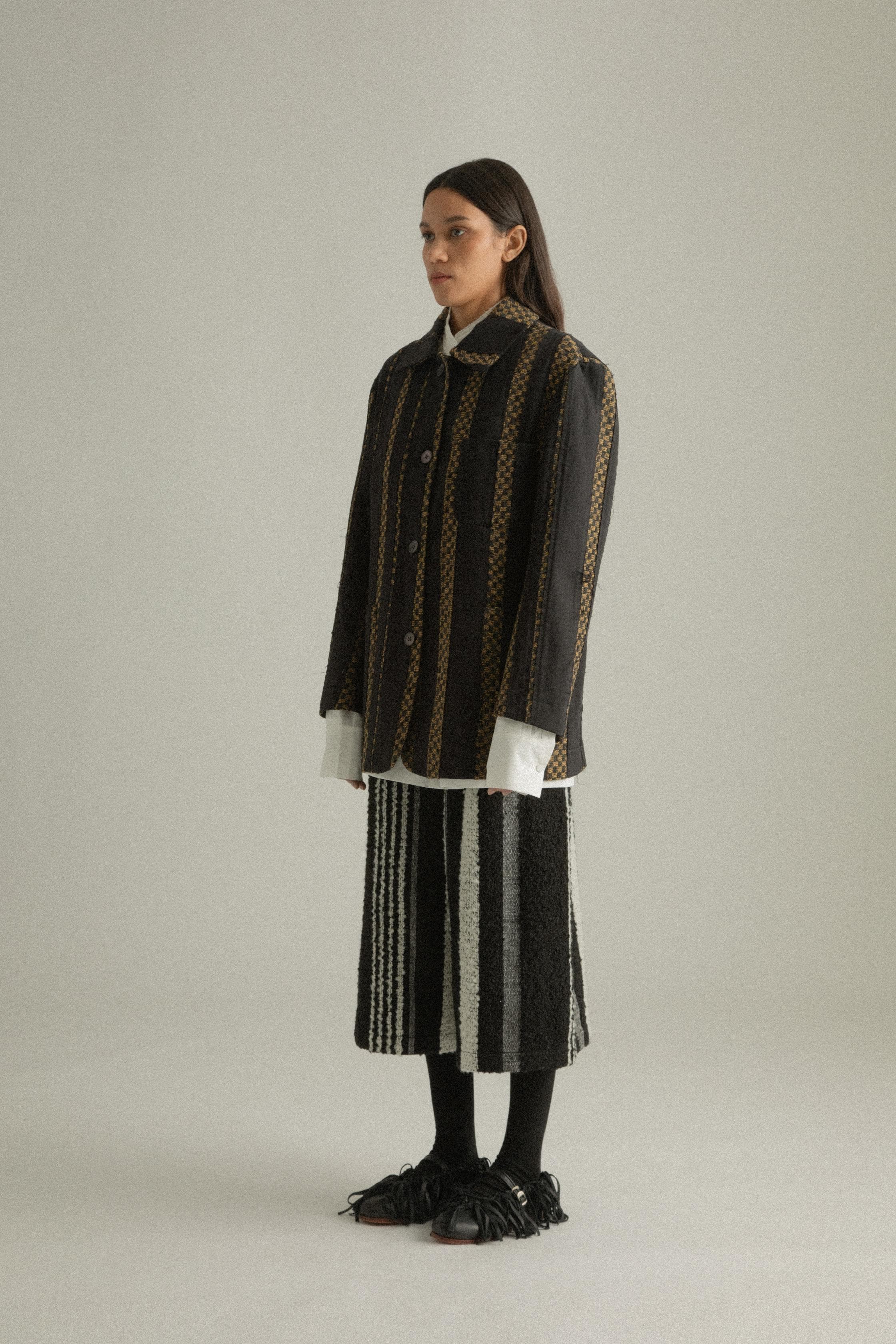 A model wearing the Nicole Jacket, featuring a classic round collar and single-button closure. The jacket is crafted from handwoven fabric in a blend of wool, cotton, and polyester with black and gold hues. Styled with DI Pants for a cohesive look. The model is shown in front, side, and back views.