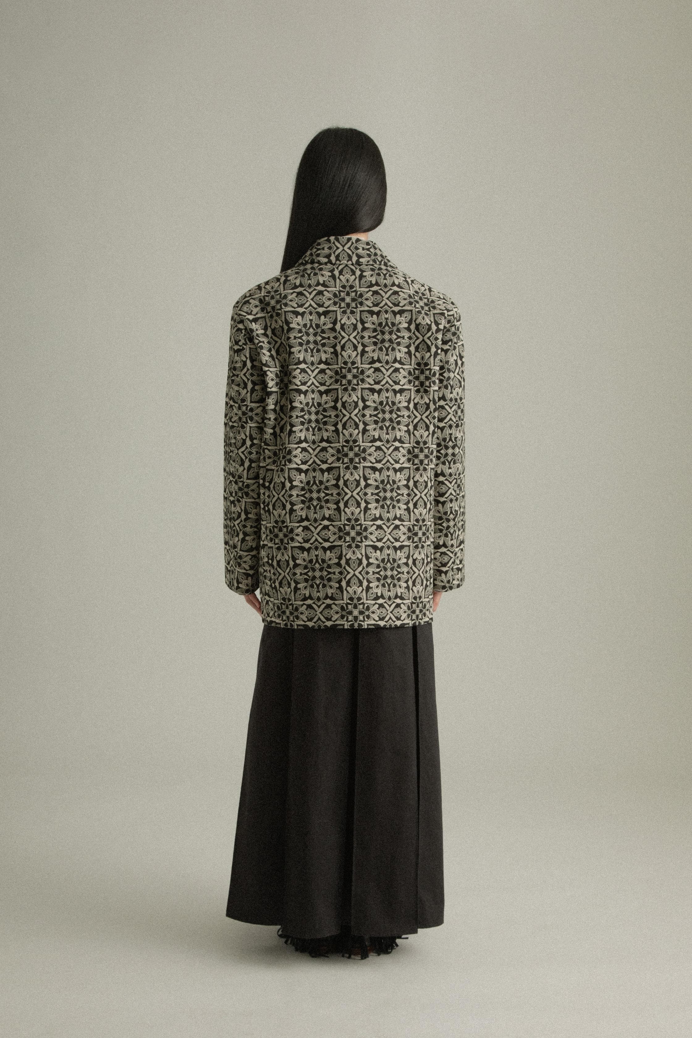 A model wearing the Nicole Jacket, featuring a classic round collar and single-button closure. Crafted from a cotton-lyocell blend with an intricate jacquard pattern inspired by mountain flora. Styled with the Rayna Pants for a balanced, textured look. The model is shown in front, side, and back views.