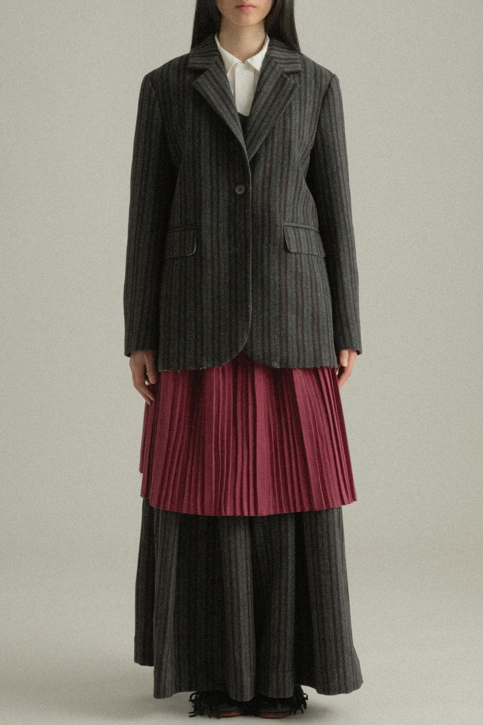 A model wearing the Nina Jacket, crafted from a wool and polyester blend with a dark grey pinstripe pattern and back pleated design. Styled with the Zuri Apron for a playful and sophisticated look. The model is shown in front, side, and back views.