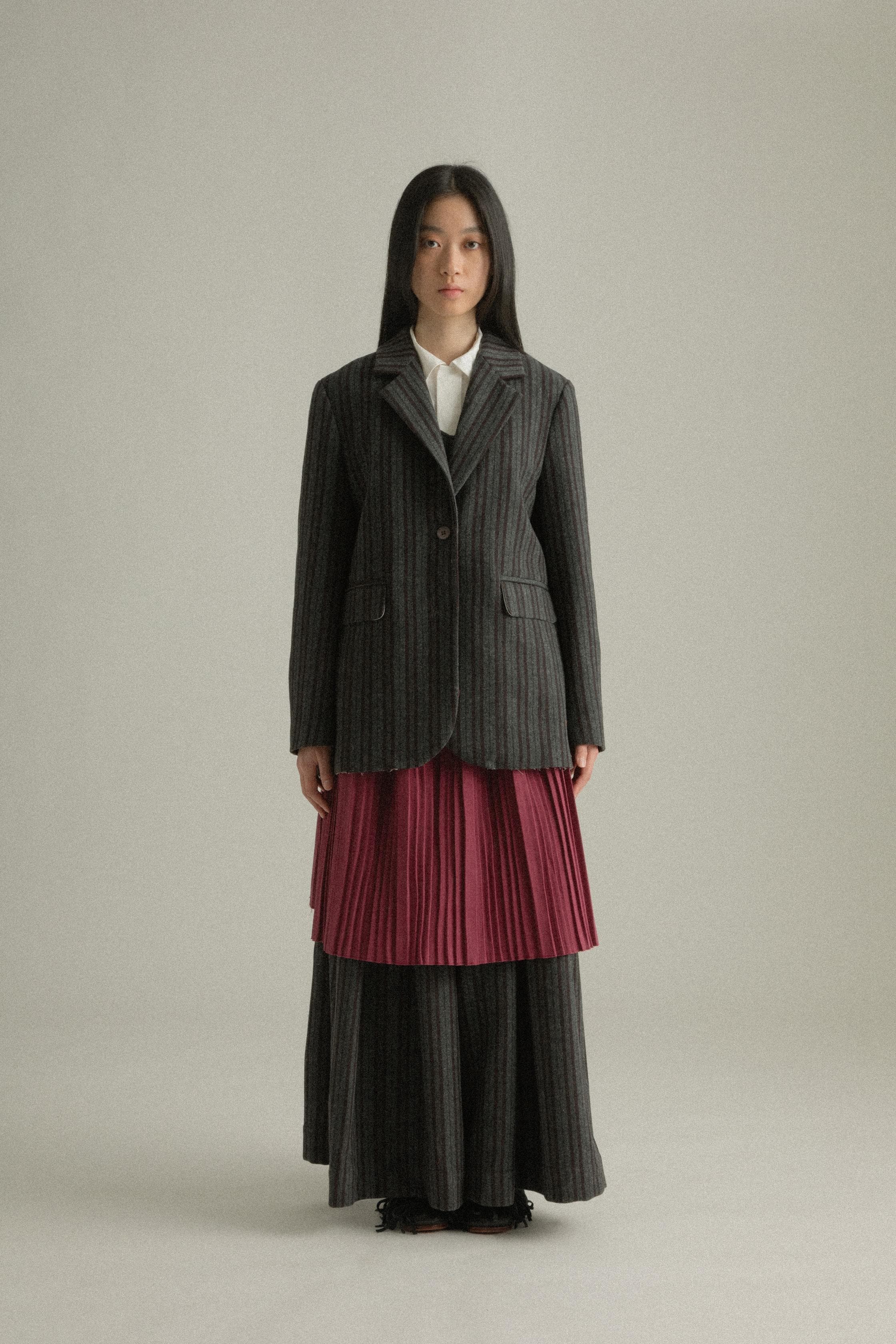 A model wearing the Nina Jacket, crafted from a wool and polyester blend with a dark grey pinstripe pattern and back pleated design. Styled with the Zuri Apron for a playful and sophisticated look. The model is shown in front, side, and back views.