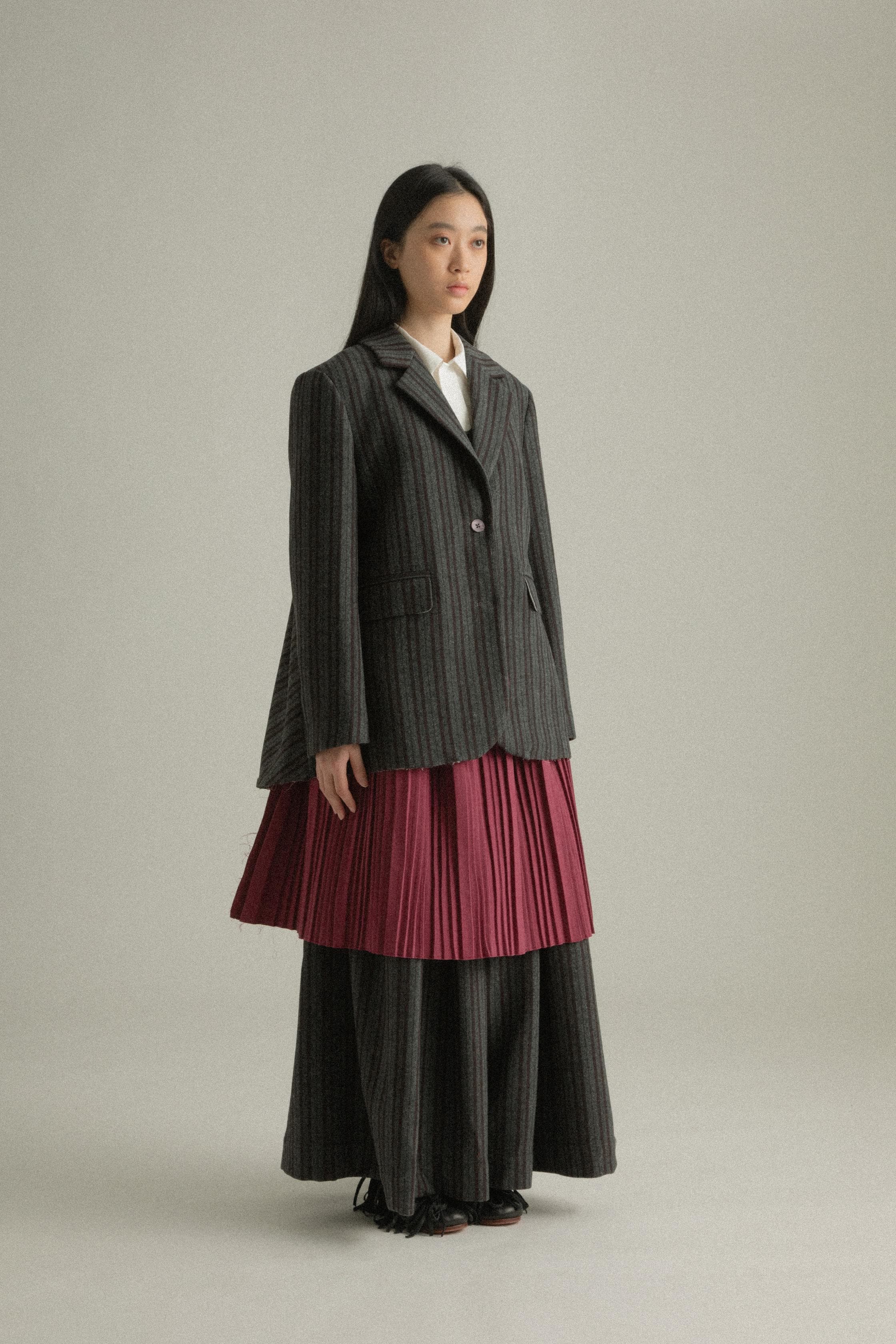 A model wearing the Nina Jacket, crafted from a wool and polyester blend with a dark grey pinstripe pattern and back pleated design. Styled with the Zuri Apron for a playful and sophisticated look. The model is shown in front, side, and back views.