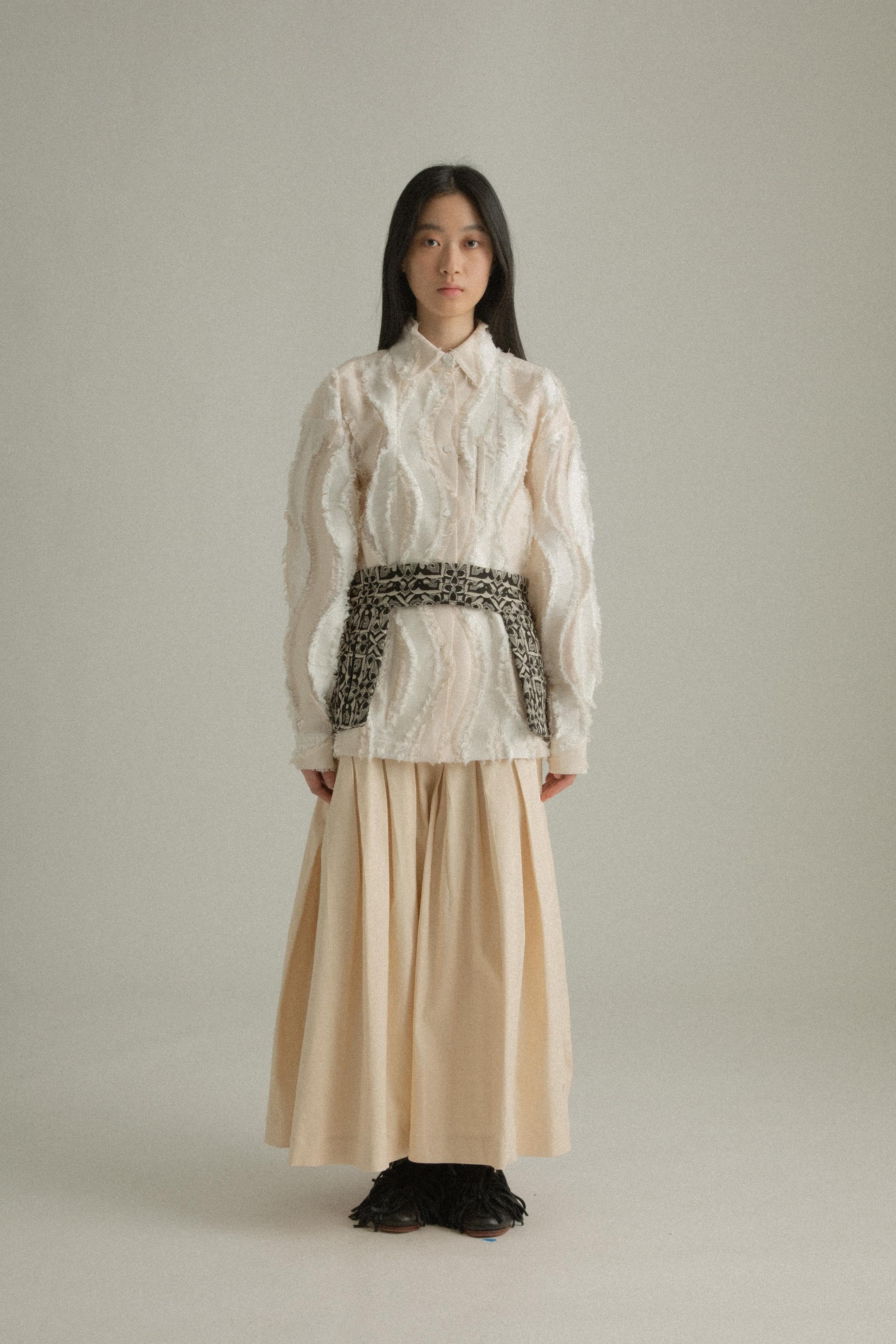 A model wearing the Percy Shirt in cream, made from Chinese cotton jacquard with a unique textural pattern. The shirt has a classic silhouette with a structured collar and concealed button placket. It is styled with the Delia Apron, which is not part of the shirt. The model is shown in front, side, and back views.
