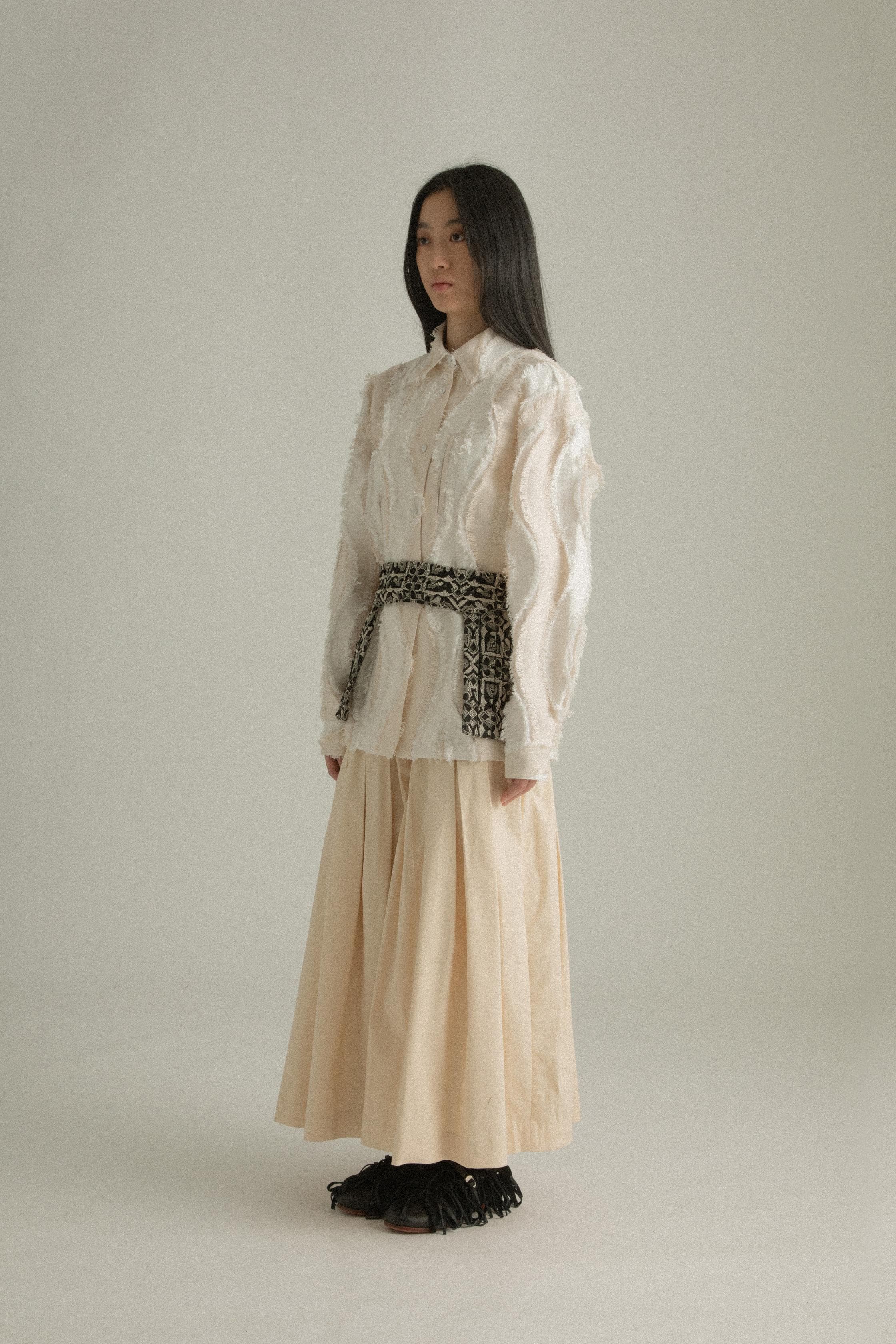 A model wearing the Percy Shirt in cream, made from Chinese cotton jacquard with a unique textural pattern. The shirt has a classic silhouette with a structured collar and concealed button placket. It is styled with the Delia Apron, which is not part of the shirt. The model is shown in front, side, and back views.
