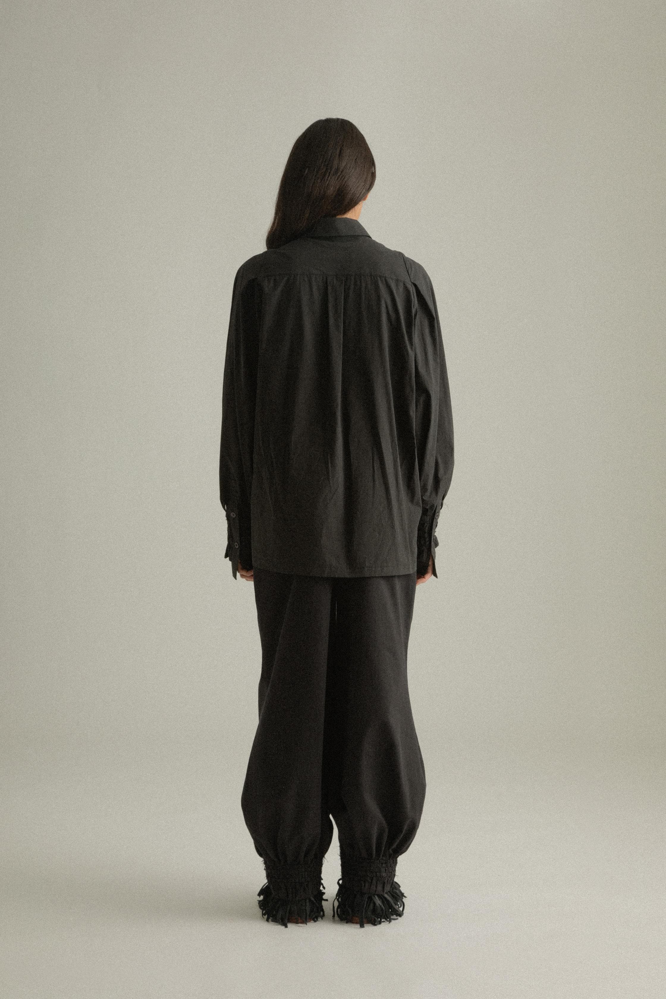 A model wearing the Qira Pants, made from organic cotton with a voluminous silhouette. The pants feature handwoven detailing at the cuffs, an elasticated waistband, and are styled with the Elias Top. The model is shown in front, side, and back views.