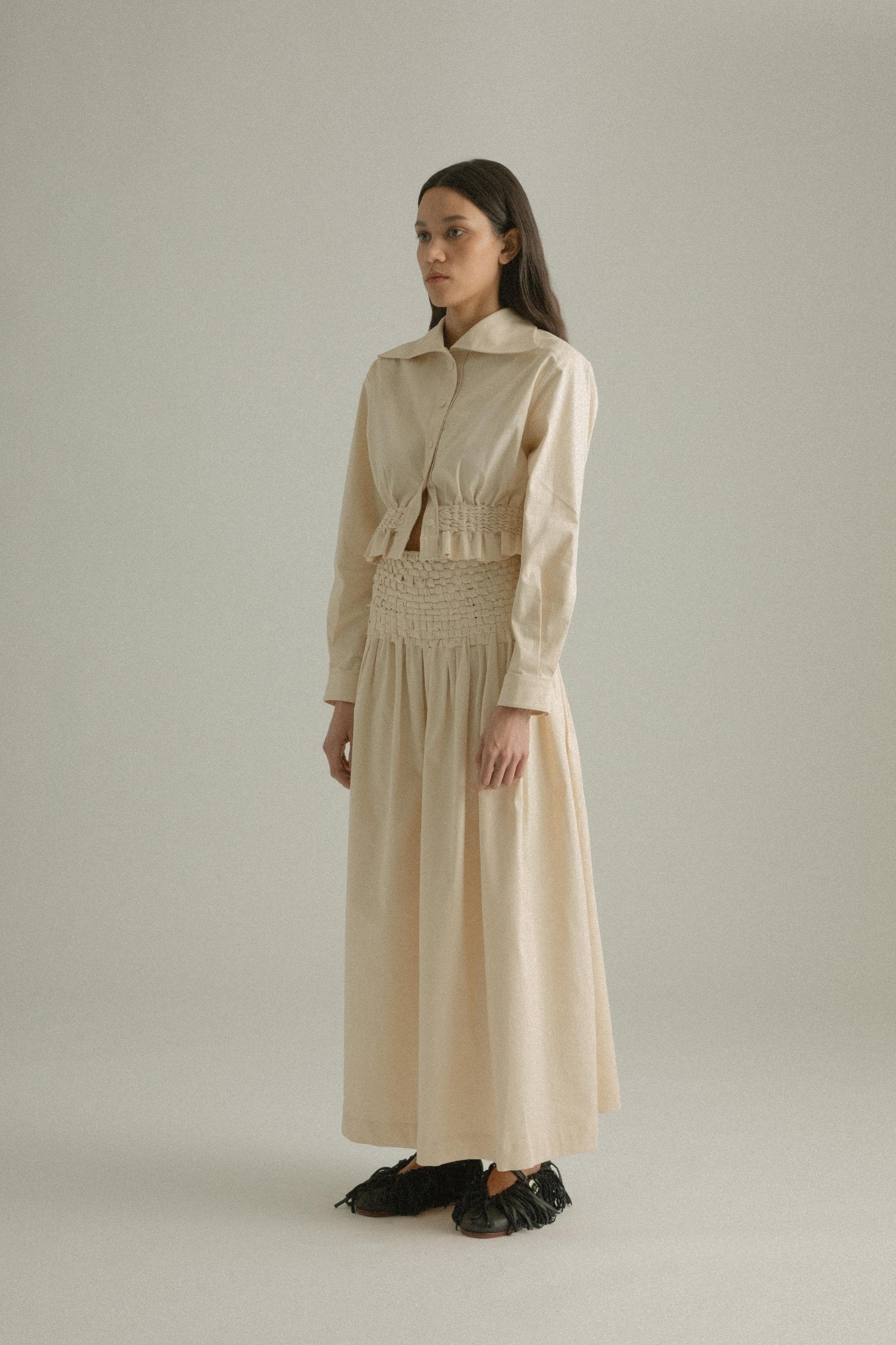 A model wearing the Rose Shirt, featuring a twist collar and hand-knit detailing at the waist. Crafted from a cotton blend with an elasticated waist, the shirt is styled with the Sylvie Skirt. The model is shown in front, side, and back views.