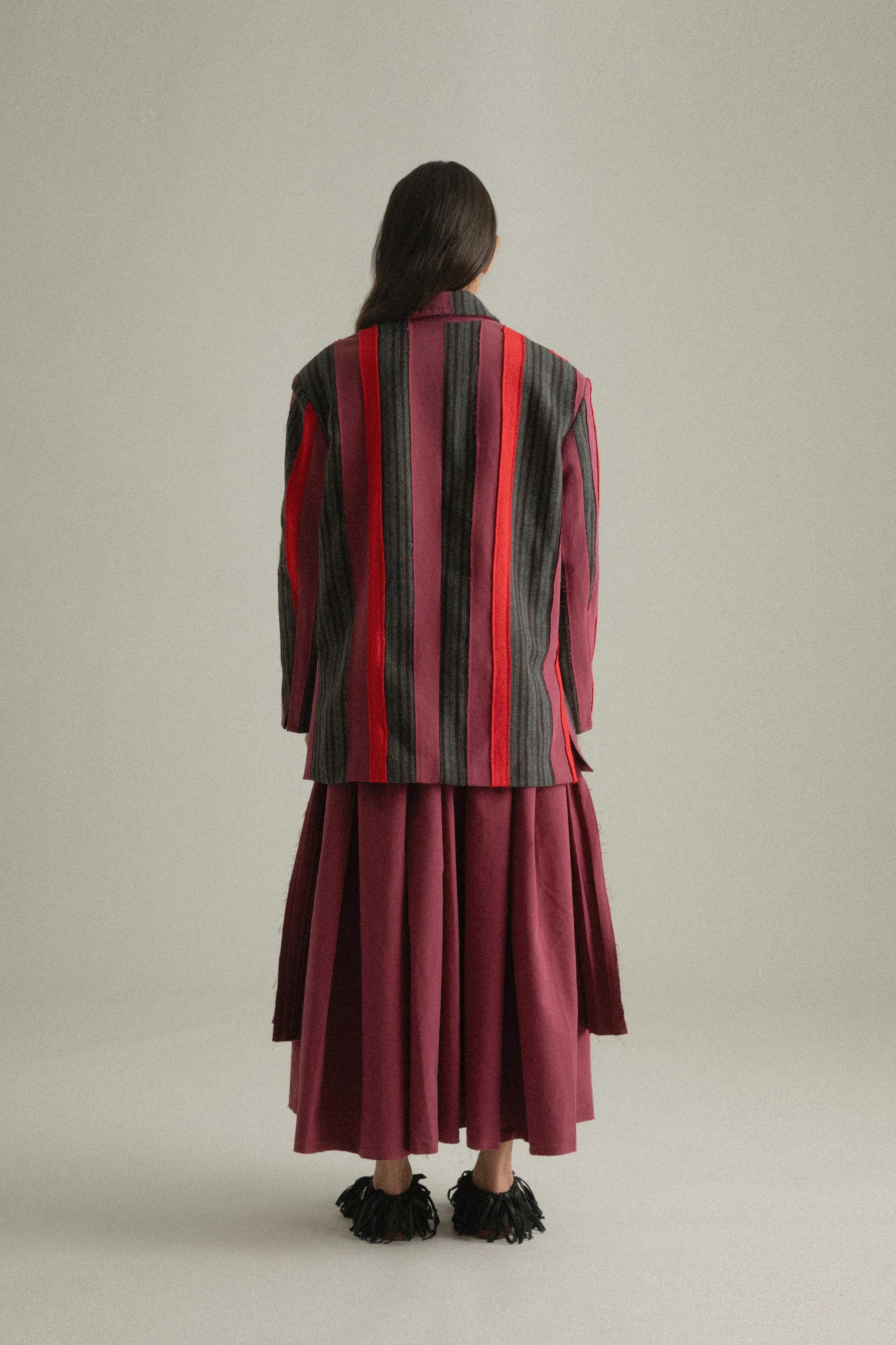 A model wearing the Thais Jacket, featuring a handwoven patchwork design in wool and cotton blends with bold colors. The jacket has a three-button closure and is styled with the Arya Dress. The model is shown in front, side, and back views.