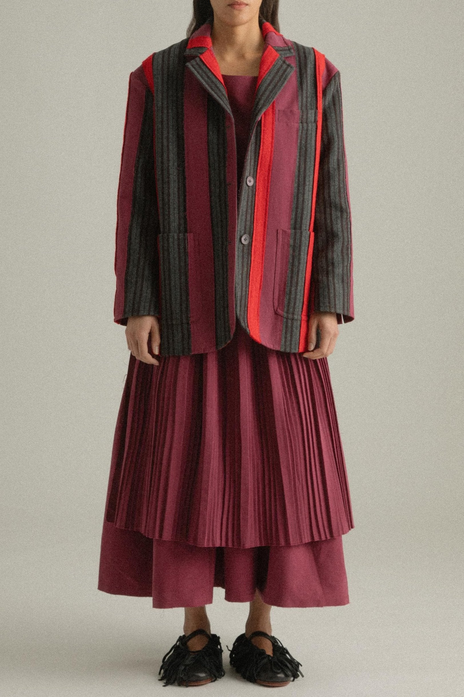 A model wearing the Thais Jacket, featuring a handwoven patchwork design in wool and cotton blends with bold colors. The jacket has a three-button closure and is styled with the Arya Dress. The model is shown in front, side, and back views.